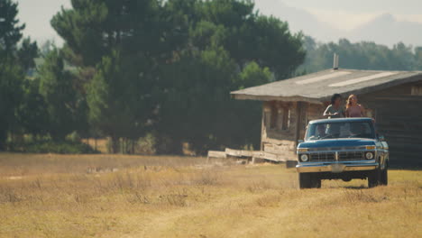 Two-Women-Standing-In-Back-Of-Pick-Up-Truck-As-Friends-Enjoy-Road-Trip-To-Countryside-Cabin