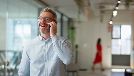 Mature-Businessman-Standing-In-Modern-Open-Plan-Office-Talking-On-Mobile-Phone