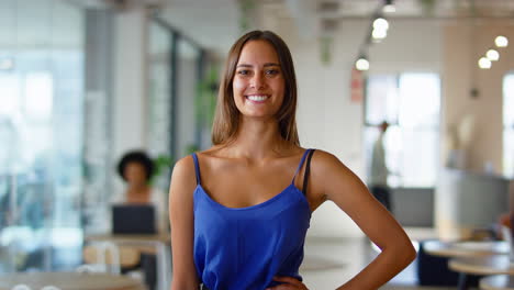Portrait-Of-Young-Smiling-Businesswoman-Standing-In-Busy-Modern-Open-Plan-Office