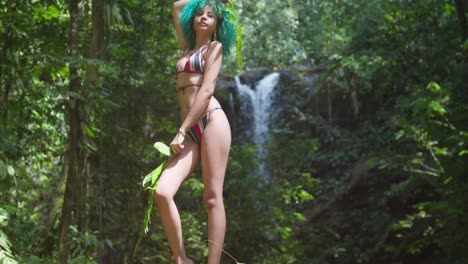 A-young-woman-stands-at-the-base-of-a-waterfall-in-a-bikini-and-green-hair