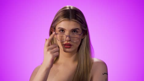 Static-shot-of-a-young-pretty-blonde-woman-putting-on-her-glasses-properly-and-looking-into-the-camera-with-an-expectant-look-in-front-of-purple-background-in-slow-motion