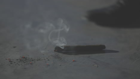 Close-up-shot-of-dropping-a-cigar-in-slowmotion-on-a-concrete-floor-with-burning-sparks-coming-off-and-smoke-rising-in-the-air-while-a-person-and-shoe-is-walking-away-in-the-background-LOG