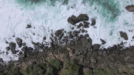 Top-down-drone-shot-of-waves-crashing-rocky-coastline-in-Indonesia-during-sunny-day