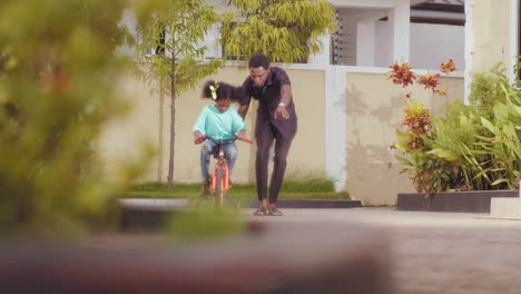 Afrikanischer-Vater-Hilft-Seiner-Tochter,-Zu-Hause-Fahrradfahren-Zu-Lernen
