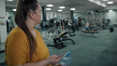 Kaukasische-Frau-Mit-Übergewicht,-Die-Im-Fitnessstudio-Ihr-Mobiltelefon-Benutzt.