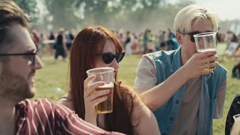 Group-of-friends-sitting-on-grass-together-at-music-festival-and-drinking-beer-while-chatting.