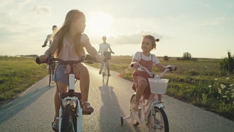 Caucasian-family-of-two-elementary-age-girls-on-foreground-and-parents-in-the-background-riding-bikes-on-village-road.