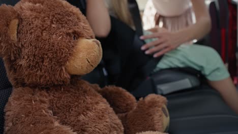 Teddy-bear-in-the-foreground-and-mother-securing-daughter-into-baby-car-seat-in-the-background.