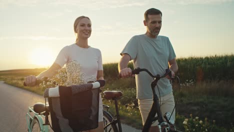 Cheerful-caucasian-couple-of-middle-age-walking-with-a-bike-on-sunset-on-village-road.