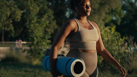 Plus-sized-African-American-woman-walking-with-exercise-mat-and-earphones-through-the-park-in-a-summer-day.