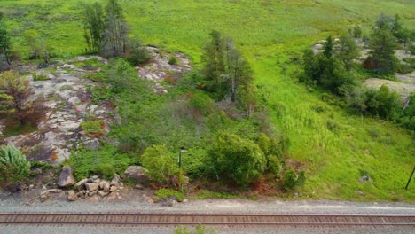Aerial-truck-pan-following-a-railway-track-passing-lush-green-natural-vegetation-which-surrounds-a-rocky-outcrop-in-Muskoka,-Ontario,-Canada