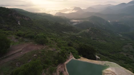 Beautiful-aerial-drone-shot-of-cliff-and-body-of-water-over-the-mountains-with-the-sunset-and-foggy-hills-in-distance