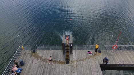 Group-of-friends-jumping-into-the-lake-from-a-wooden-pier,-Aerial-View