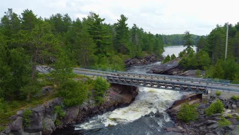 Aerial-drone-shot-following-a-fast-flowing-river-surrounded-by-trees,-passing-above-a-motor-vehicle-bridge-and-into-the-Magnetawan-River,-Ontario,-Canada