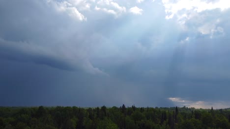 An-aerial-drone-shot-of-sunrays-shining-through-thick-dramatic-storm-clouds-building-in-the-atmosphere-over-a-forest-in-Muskoka,-Canada