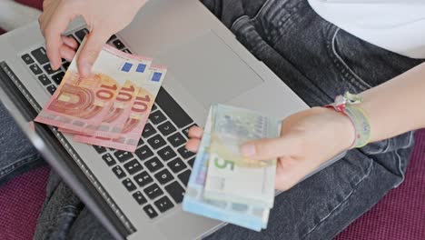 Hands-of-young-woman-counting-euros-on-laptop,-view-from-above