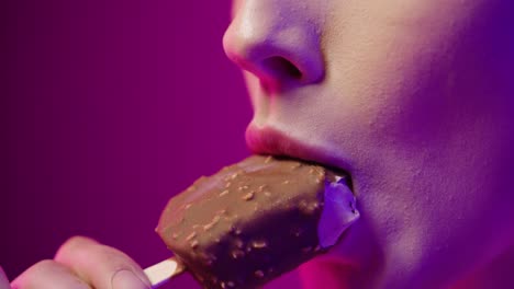 Close-up-shot-of-a-young-woman-with-beautiful-red-lips-while-eating-a-vegan-chocolate-ice-cream-on-a-stick-and-enjoying-the-taste-in-front-of-purple-background-in-slow-motion