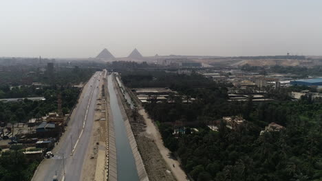 Aerial-Shot-Track-out-and-rising-up-for-The-Pyramids-of-Egypt-in-Giza-in-the-background-of-a-branch-of-the-River-Nile-in-the-foreground-Maryotya-branch-showing-greenery-in-the-surroundings