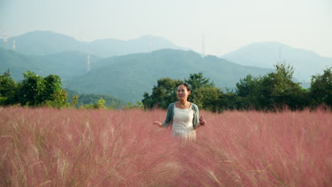 Eine-Asiatische-Koreanerin-Geht-In-Zeitlupe-Durch-Ein-Rosafarbenes,-Hohes-Grasfeld-Mit-Malerischem-Gebirgshintergrund-Auf-Der-Kräuterinselfarm-Im-Pocheon-Hochland-Auf-Die-Kamera-Zu