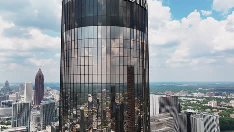 Aerial-descending-shot-of-mirror-facade-of-modern-cylindrical-skyscraper-reflecting-surrounding-buildings-and-clouds-in-sky