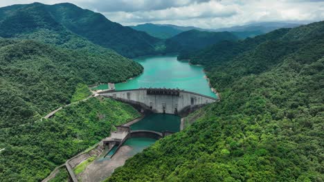 Aerial-backwards-shot-of-Feitsui-Dam-surrounded-by-tropical-mountain-landscape-during-cloudy-day