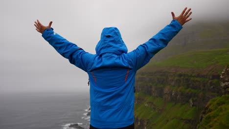 Man-raising-arms-while-enjoying-view-of-foggy-mountain-in-Gasadalur,-Faroe-Islands