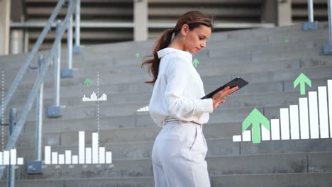 Successful-Businesswoman-working-on-tablet-stands-between-various-digital-graphs,-positive-arrows-and-bar-charts-on-stairs-of-office-building