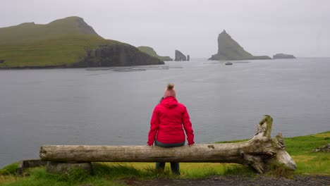 Woman-sitting-on-log-looking-at-Drangarnir-and-Tindholmur-in-Vagar,-Faroe-Islands
