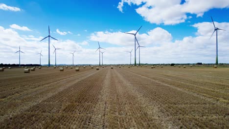 Oben-Dreht-Sich-Eine-Reihe-Von-Windkraftanlagen-Anmutig-Auf-Dem-Kürzlich-Abgeernteten-Feld-Eines-Bauern-In-Lincolnshire,-Vorne-Mit-Goldenen-Heuballen