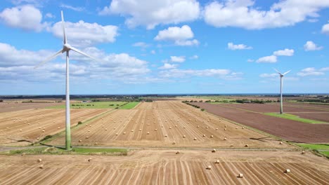 The-aerial-footage-reveals-a-picturesque-sight—a-row-of-wind-turbines-spinning-gracefully-within-a-Lincolnshire-farmer's-newly-harvested-field,-where-golden-hay-bales-add-rustic-charm