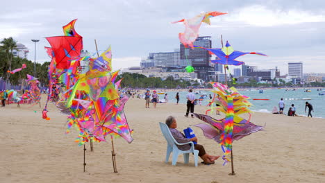 Colorful-kites-are-displayed-on-bamboo-poles-at-the-foreground,-and-in-the-background-beachgoers-are-swimming,-sailing,-and-strolling-at-the-beach-in-Pattaya-at-Chonburi-province-in-Thailand