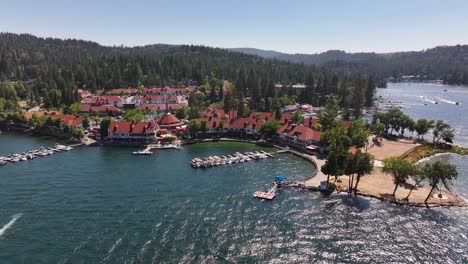 boats-and-yachts-leaving-dock-in-front-of-Lake-Arrowhead-Village-waterfront-in-California-on-a-bright-summer-day-AERIAL-TRUCKING-PAN
