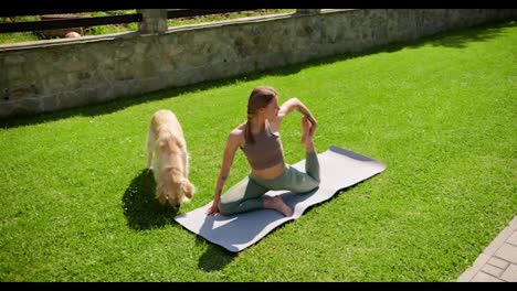 A-blonde-girl-in-a-special-sports-summer-uniform-does-yoga-on-a-gray-rug-on-the-lawn,-her-light-colored-dog-walks-beside-her