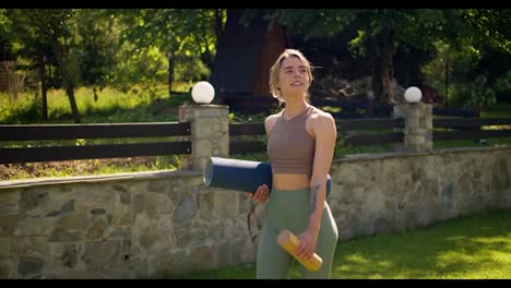 A-young-blonde-girl-in-a-sports-summer-uniform-walks-along-a-green-lawn-along-a-stone-fence-and-holds-a-karimat-and-a-bottle-of-water-in-her-hands.-Sports-and-yoga-in-nature