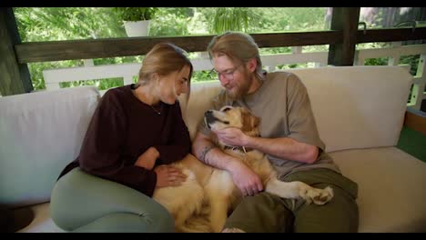 A-blonde-girl-in-a-red-sweater-and-a-blonde-guy-in-glasses-with-a-beard-in-a-light-brown-T-shirt-are-stroking-their-light-colored-dog-on-a-sofa-in-nature-in-the-gazebo