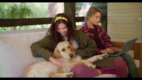A-blonde-girl-in-a-red-plaid-shirt-works-on-a-laptop,-and-a-brunette-girl-in-a-green-jacket-strokes-a-light-colored-dog-in-a-gazebo-in-nature