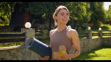A-blonde-girl-in-summer-sportswear-walks-along-a-green-lawn-and-prepares-for-yoga.-The-girl-carries-a-special-yoga-mat-and-a-bottle-of-water-in-her-hands
