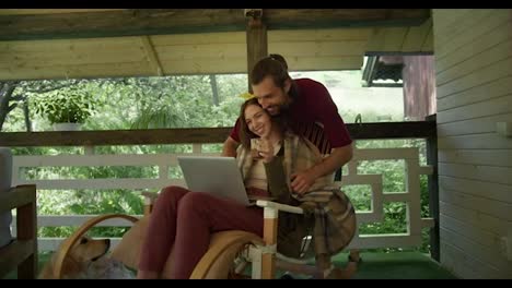 A-brunette-guy-in-a-red-T-shirt-envelops-a-girl-while-she-sits-on-an-armchair,-looks-at-a-laptop-screen-and-drinks-tea-n-the-gazebo-against-the-backdrop-of-the-forest.-The-manifestation-of-care-of-a-young-guy-is-taken-to-a-girl
