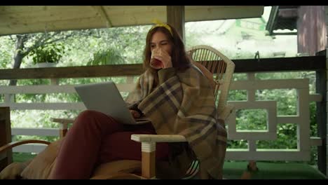 A-brunette-girl-sits-on-an-armchair-in-a-plaid-with-tea-and-looks-at-the-screen-of-a-laptop-that-she-has-on-her-knees-in-the-gazebo-against-the-backdrop-of-a-green-forest