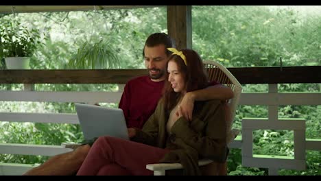 A-brunette-guy-in-a-red-blouse-and-a-brunette-girl-in-a-green-blouse-are-sitting-on-an-armchair,-talking-and-looking-at-something-in-a-laptop-in-the-gazebo-against-the-backdrop-of-a-green-forest