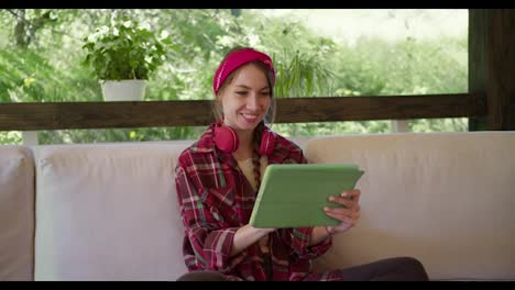 A-blonde-girl-in-a-red-plaid-shirt-with-red-headphones-and-a-red-bandage-sits-and-works-on-a-green-tablet-on-a-sofa-in-a-gazebo-in-nature