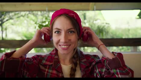 shooting-close-up-a-girl-in-a-red-plaid-shirt-puts-on-red-wireless-headphones-listening-to-music-and-shaking-her-head-Sitting-on-a-sofa-in-a-gazebo-in-nature