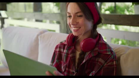 Close-up-shot:-a-girl-in-red-clothes-and-red-headphones-sits-on-a-sofa-and-works-on-a-green-tablet-in-a-gazebo-in-nature