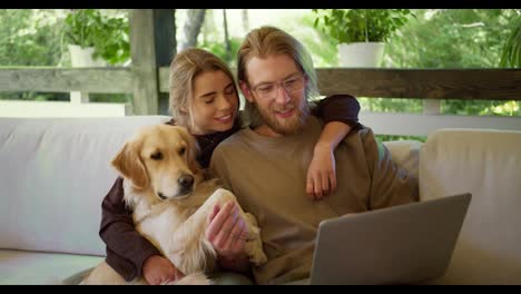 A-blond-girl-and-a-blond-guy-with-a-beard-in-glasses-are-looking-at-something-funny-on-a-laptop-and-laughing-next-to-them-their-dog-is-sitting.-Leisure-in-the-gazebo-in-nature