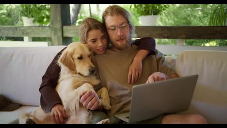 A-happy-family-of-a-guy-and-a-blonde-girl,-together-with-their-dog,-are-considering-goods-in-an-online-store-on-their-laptop.-Sitting-on-a-sofa-in-a-gazebo-in-nature