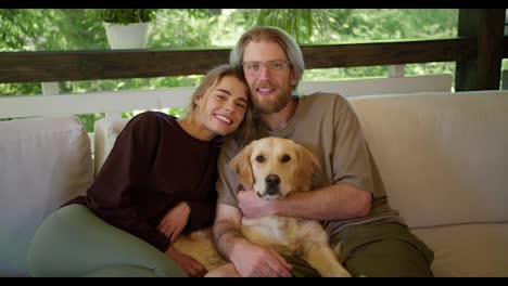 Portrait-of-a-happy-couple-of-a-guy-and-a-blonde-girl-together-with-their-dog.-A-guy-with-glasses-with-a-beard-in-a-light-brown-T-shirt-a-girl-in-a-dark-red-T-shirt,-they-are-sitting-with-their-light-colored-dog-on-a-sofa-in-a-gazebo-in-nature