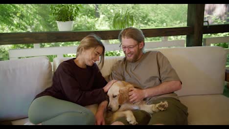 A-blond-girl-in-a-red-sweater-and-a-blond-guy-in-glasses-with-a-beard-in-a-light-brown-T-shirt-are-sitting-on-a-devan-and-stroking-their-big-light-colored-dog-on-a-devan-in-a-gazebo-in-nature