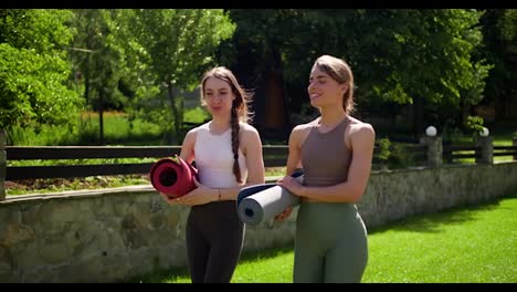 2-blonde-girls-in-summer-sportswear-go-to-yoga-classes-and-carry-special-mats.-Preparing-for-a-yoga-class-in-a-green-park-in-nature