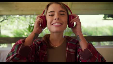 Blonde-girl-in-a-red-plaid-shirt-puts-on-red-wireless-headphones-smiles-looks-at-the-camera-and-shakes-her-head-Sitting-on-a-sofa-in-a-gazebo-in-nature