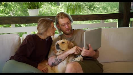 A-blond-girl-and-a-blond-guy-with-a-beard-in-glasses-are-looking-at-something-on-a-laptop,-a-dog-is-sitting-next-to-them,-which-the-girl-is-stroking.-Leisure-on-the-sofa-in-the-gazebo-in-nature